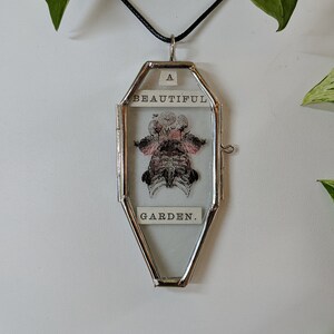 May include: A silver locket necklace with a glass insert featuring a pressed flower and the text "A Beautiful Garden."