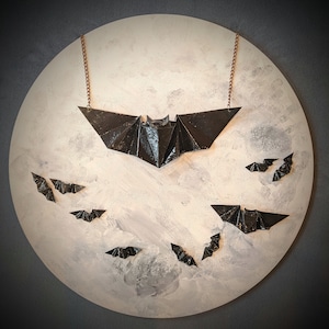 May include: A black origami bat necklace with a chain and a white background. There are smaller black origami bats scattered around the larger bat.