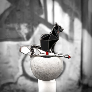 Pode incluir: Um pin de gato geométrico preto com boca vermelha e contorno prateado, pousado numa vassoura branca e prateada. O pin está sobre um fundo em tons de cinza, em cima de uma esfera de pedra cinzenta.