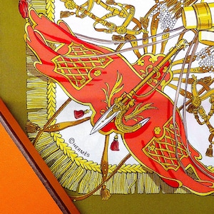 May include: A silk scarf with a detailed design featuring red, gold, and white elements. The scarf is in an orange box with the brand name "HERMES" visible. The scarf has a military theme.