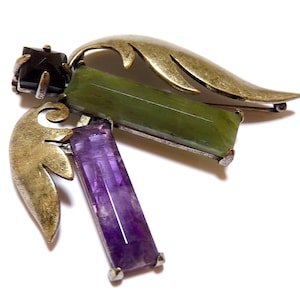 May include: A gold-tone brooch with a green, purple, and black gemstone design. The brooch features a stylized wing shape with intricate details.
