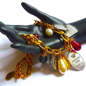 Puede incluir: Un brazalete de cadena dorada con m&uacute;ltiples dijes, incluyendo una cuenta de vidrio roja y verde, una perla y un dije de plata con el texto "JACQUES FATH PARIS".