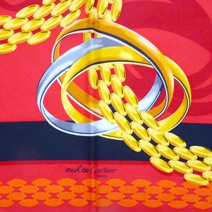 May include: A red, black, and orange silk scarf with a gold chain and two interlocking rings. The rings are silver and gold. The scarf has the words 'must de Cartier PARIS' printed on it.