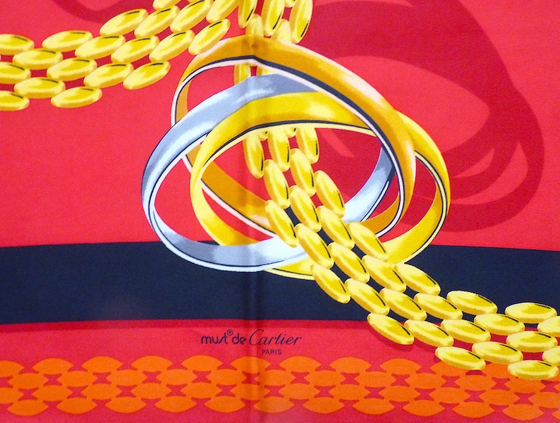 May include: A red, black, and orange silk scarf with a gold chain and two interlocking rings. The rings are silver and gold. The scarf has the words 'must de Cartier PARIS' printed on it.