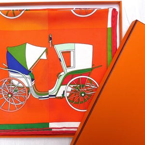 May include: A vintage-style horse-drawn carriage with a white top and green accents, printed on a bright orange silk scarf. The scarf is folded and displayed in a matching orange box.