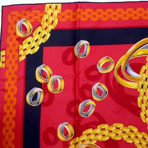 May include: Red silk scarf with a black border and a pattern of gold chains and silver rings. The scarf has a repeating pattern of gold chains and silver rings. The chains are linked together and the rings are overlapping. The background is red and the border is black.