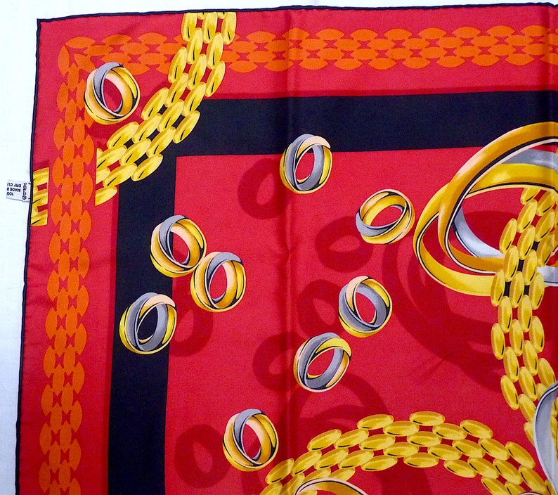 May include: Red silk scarf with a black border and a pattern of gold chains and silver rings. The scarf has a repeating pattern of gold chains and silver rings. The chains are linked together and the rings are overlapping. The background is red and the border is black.
