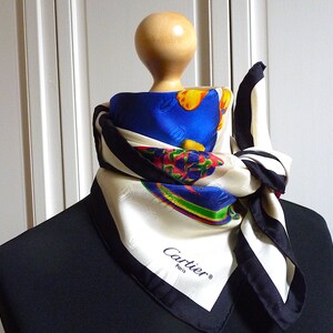 May include: A white silk scarf with a black and white border. The scarf has a colorful design with blue, yellow, red, and green. The scarf is tied around a mannequin's neck and has the word 'Cartier' and 'Paris' printed on it.