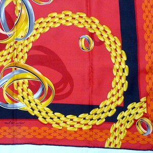 May include: A red silk scarf with a black border and a gold chain pattern. The chain pattern is made up of interlocking rings. The scarf also features a silver ring pattern. The scarf has the text 'must de Cartier Paris' in the lower left corner.
