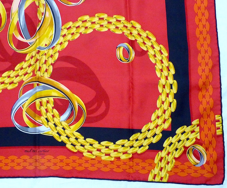 May include: A red silk scarf with a black border and a gold chain pattern. The chain pattern is made up of interlocking rings. The scarf also features a silver ring pattern. The scarf has the text 'must de Cartier Paris' in the lower left corner.