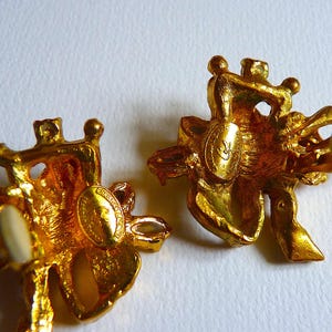 K&ouml;nnte beinhalten: Ein Paar goldfarbene Ohrringe mit Blumenmuster. Die Ohrringe haben einen wei&szlig;en Stein in der Mitte jeder Blume. Die Ohrringe sind auf der R&uuml;ckseite mit dem Buchstaben "C" gekennzeichnet.