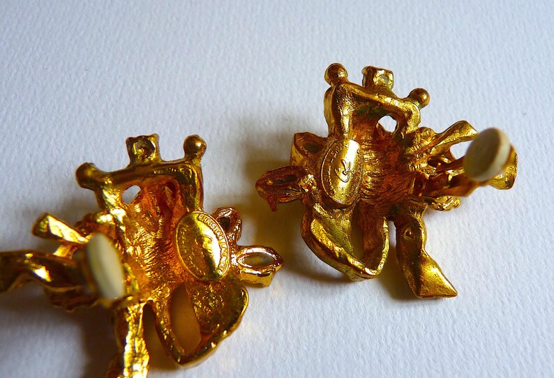 K&ouml;nnte beinhalten: Ein Paar goldfarbene Ohrringe mit Blumenmuster. Die Ohrringe haben einen wei&szlig;en Stein in der Mitte jeder Blume. Die Ohrringe sind auf der R&uuml;ckseite mit dem Buchstaben "C" gekennzeichnet.