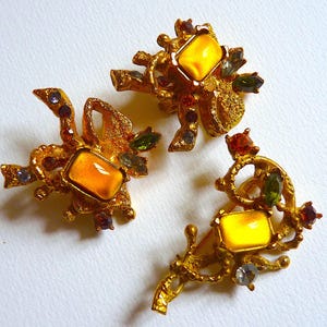 K&ouml;nnte beinhalten: Drei goldfarbene Broschen mit filigranen Designs und gelben Glassteinen. Jede Brosche hat ein anderes Design mit verschiedenen farbigen Steinen.