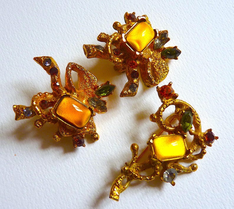 K&ouml;nnte beinhalten: Drei goldfarbene Broschen mit filigranen Designs und gelben Glassteinen. Jede Brosche hat ein anderes Design mit verschiedenen farbigen Steinen.