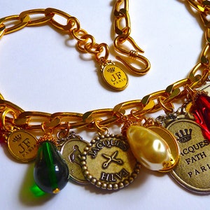 Puede incluir: Un brazalete de cadena dorada con m&uacute;ltiples dijes, incluyendo una perla, cuentas de vidrio rojas y verdes, y dijes de metal con el texto "JF Paris", "Jacques Fath Paris" y "Hilfiger".