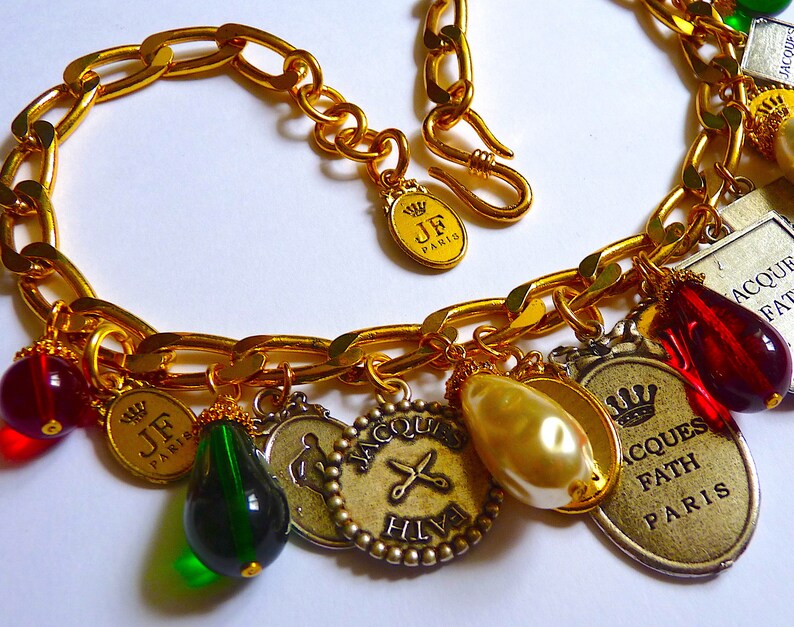 Puede incluir: Un brazalete de cadena dorada con m&uacute;ltiples dijes, incluyendo una perla, cuentas de vidrio rojas y verdes, y dijes de metal con el texto "JF Paris", "Jacques Fath Paris" y "Hilfiger".