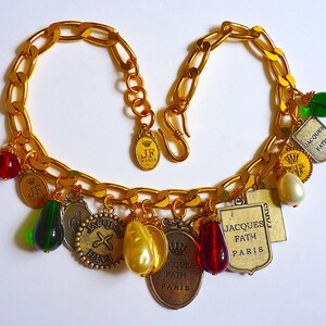 Puede incluir: Un collar de cadena dorada con m&uacute;ltiples dijes, incluyendo un par de tijeras, una corona y varias etiquetas peque&ntilde;as con el texto "Jacques Fath Paris". Los dijes est&aacute;n unidos a la cadena con peque&ntilde;os anillos dorados. El collar est&aacute; sobre un fondo blanco.