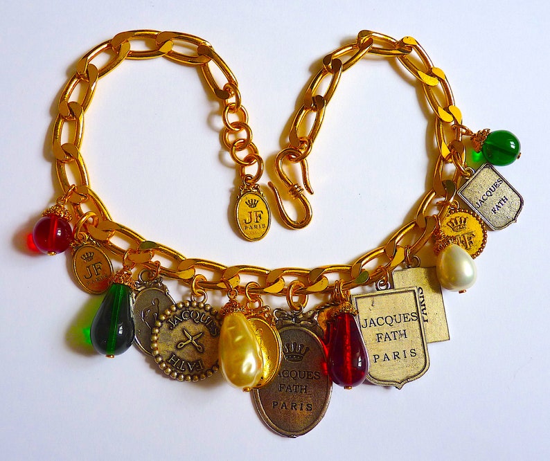Puede incluir: Un collar de cadena dorada con m&uacute;ltiples dijes, incluyendo un par de tijeras, una corona y varias etiquetas peque&ntilde;as con el texto "Jacques Fath Paris". Los dijes est&aacute;n unidos a la cadena con peque&ntilde;os anillos dorados. El collar est&aacute; sobre un fondo blanco.