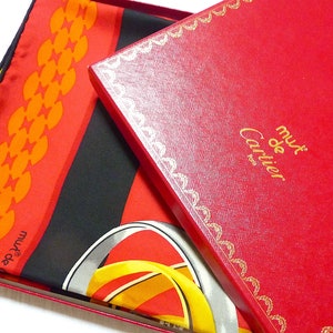 May include: A red and black silk scarf with a gold and yellow design. The scarf is folded and placed on top of a red box with gold trim. The box has the words 'must de Cartier' printed on it in gold letters.