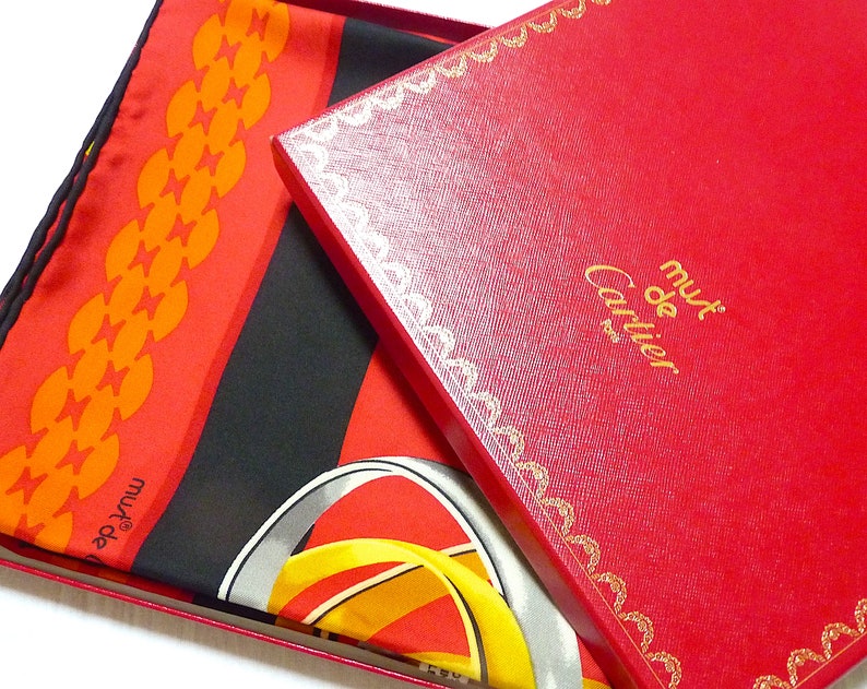 May include: A red and black silk scarf with a gold and yellow design. The scarf is folded and placed on top of a red box with gold trim. The box has the words 'must de Cartier' printed on it in gold letters.
