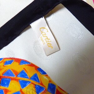 May include: A white silk scarf with a black trim and a yellow tag that says 'Cartier' in gold lettering. The scarf has a repeating pattern of colorful, abstract shapes.