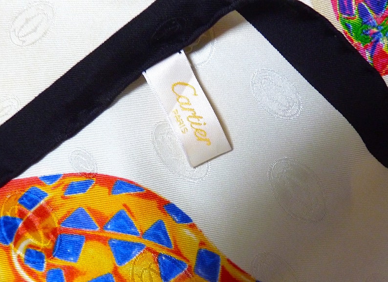 May include: A white silk scarf with a black trim and a yellow tag that says 'Cartier' in gold lettering. The scarf has a repeating pattern of colorful, abstract shapes.