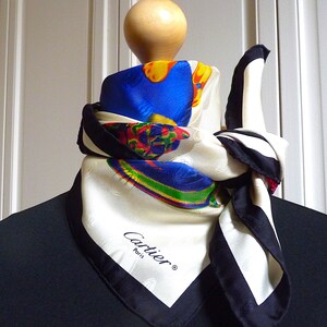 May include: A white silk scarf with a black border and a colorful abstract design. The scarf is tied around the neck of a mannequin. The scarf has the text 'Cartier Paris' printed on it.