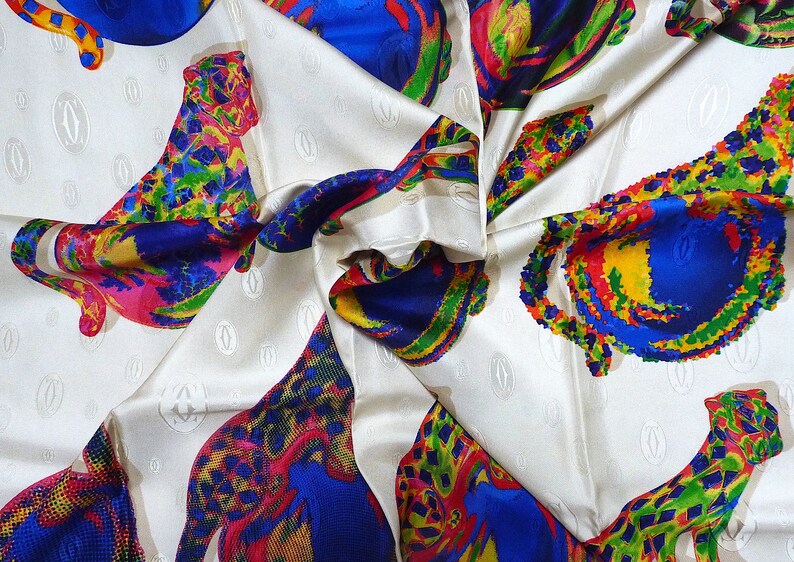 May include: A white silk scarf with a repeating pattern of colorful leopards. The leopards are made up of bright colors including red, yellow, blue, green, and pink. The scarf has a subtle embossed pattern of the word 'Cartier' repeated throughout.