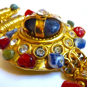 Könnte beinhalten: Eine goldfarbene Halskette mit einem großen, verzierten Anhänger, der einen blauen Stein zeigt, der von kleineren Steinen und Strasssteinen umgeben ist. Der Anhänger ist an einer Kette mit mehreren goldfarbenen Gliedern befestigt.