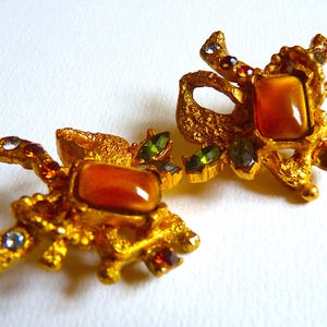 K&ouml;nnte beinhalten: Ein Paar goldfarbene Ohrringe mit einem gro&szlig;en, orangebraunen Stein, der in einem strukturierten, verzierten Design gefasst ist. Die Ohrringe sind mit kleineren, gr&uuml;nen und roten Steinen verziert.