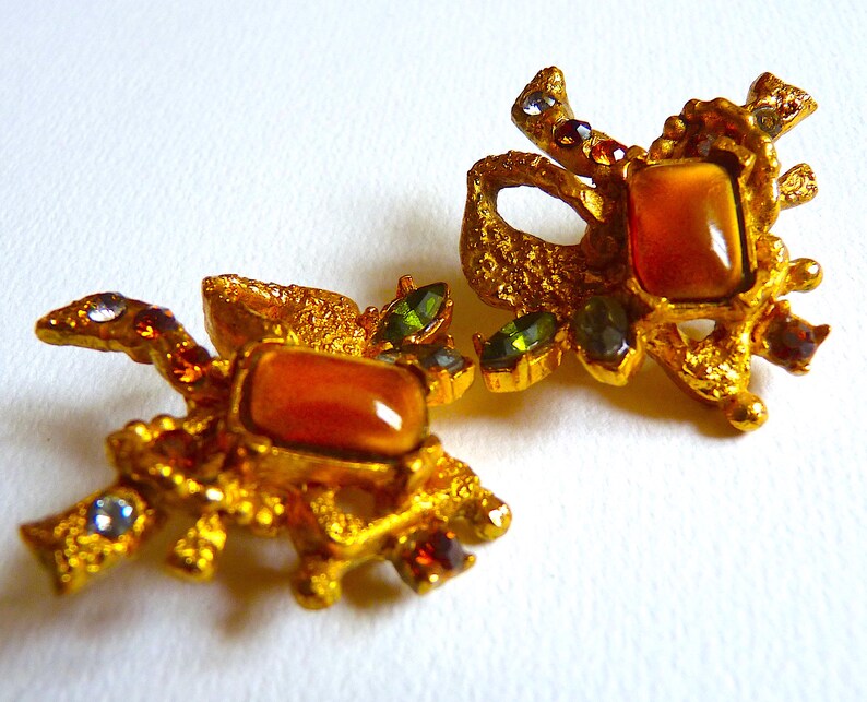 K&ouml;nnte beinhalten: Ein Paar goldfarbene Ohrringe mit einem gro&szlig;en, orangebraunen Stein, der in einem strukturierten, verzierten Design gefasst ist. Die Ohrringe sind mit kleineren, gr&uuml;nen und roten Steinen verziert.
