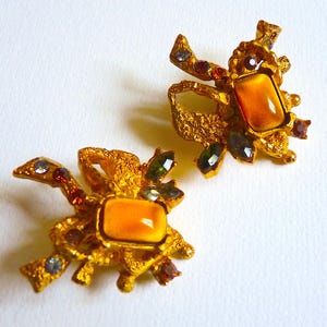 K&ouml;nnte beinhalten: Ein Paar goldfarbene Ohrringe mit strukturiertem Design. Jeder Ohrring hat einen gro&szlig;en, rechteckigen, orangefarbenen Stein, der von kleineren, mehrfarbigen Steinen umgeben ist.