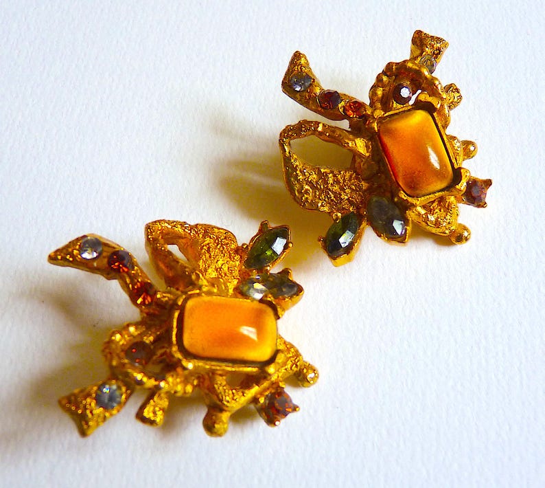 K&ouml;nnte beinhalten: Ein Paar goldfarbene Ohrringe mit strukturiertem Design. Jeder Ohrring hat einen gro&szlig;en, rechteckigen, orangefarbenen Stein, der von kleineren, mehrfarbigen Steinen umgeben ist.