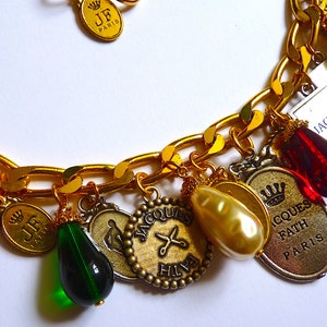 Puede incluir: Un brazalete de oro con varios dijes, incluyendo una cuenta de vidrio roja, una cuenta de vidrio verde, una perla y un dije con el texto "JACQUES FATH PARIS".