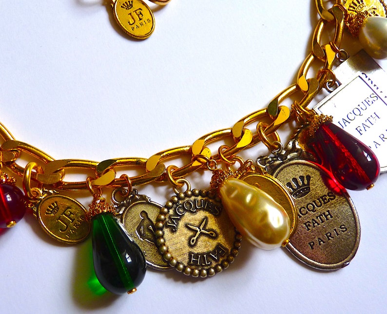 Puede incluir: Un brazalete de oro con varios dijes, incluyendo una cuenta de vidrio roja, una cuenta de vidrio verde, una perla y un dije con el texto "JACQUES FATH PARIS".