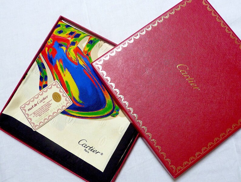 May include: A white silk scarf with a colorful abstract design. The scarf is folded and displayed in a red box with gold trim. The box has the word 'Cartier' printed in gold on the front. The scarf has a tag attached to it that says 'must de Cartier' and 'Paris'. The scarf is a luxury item.