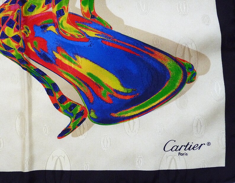 May include: A white silk scarf with a colorful abstract design of a shoe. The scarf has a black border and the words 'Cartier® Paris' printed in the bottom right corner.