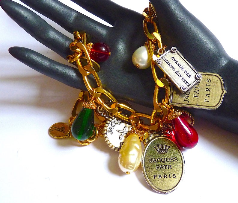 Puede incluir: Un brazalete de oro con m&uacute;ltiples dijes, incluyendo una l&aacute;grima de vidrio verde, una l&aacute;grima de vidrio rojo, una perla blanca y un dije de oro con el texto "Jacques Fath Paris".