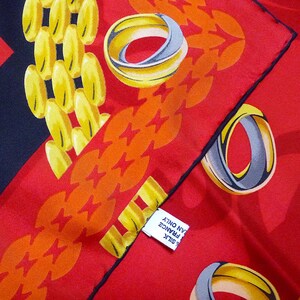 May include: A red silk scarf with a black border. The scarf has a pattern of gold chains and rings. The text on the tag reads 'SILK', 'MADE IN FRANCE', and 'CLEAN ONLY'. 
