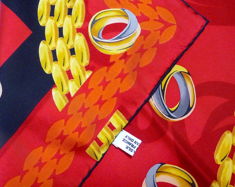 May include: A red silk scarf with a black border. The scarf has a pattern of gold chains and rings. The text on the tag reads 'SILK', 'MADE IN FRANCE', and 'CLEAN ONLY'. 
