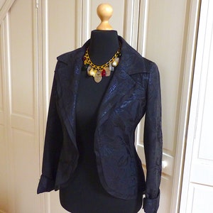 Puede incluir: Una chaqueta negra con estampado floral, con un collar de cadena dorada con colgantes. La chaqueta tiene un cuello chal y mangas largas.