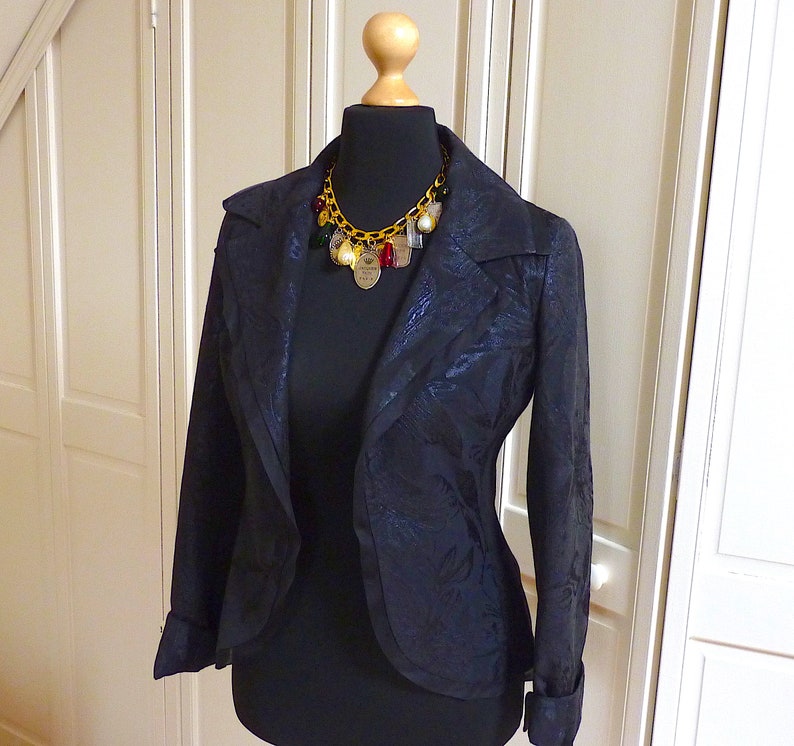 Puede incluir: Una chaqueta negra con estampado floral, con un collar de cadena dorada con colgantes. La chaqueta tiene un cuello chal y mangas largas.