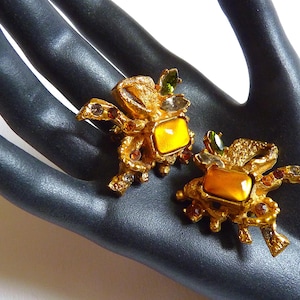 K&ouml;nnte beinhalten: Ein Paar goldfarbene Ohrringe mit einem Krabbenmotiv. Jeder Ohrring hat einen gro&szlig;en, gelb-orangen Edelstein, der von kleineren, mehrfarbigen Edelsteinen umgeben ist.