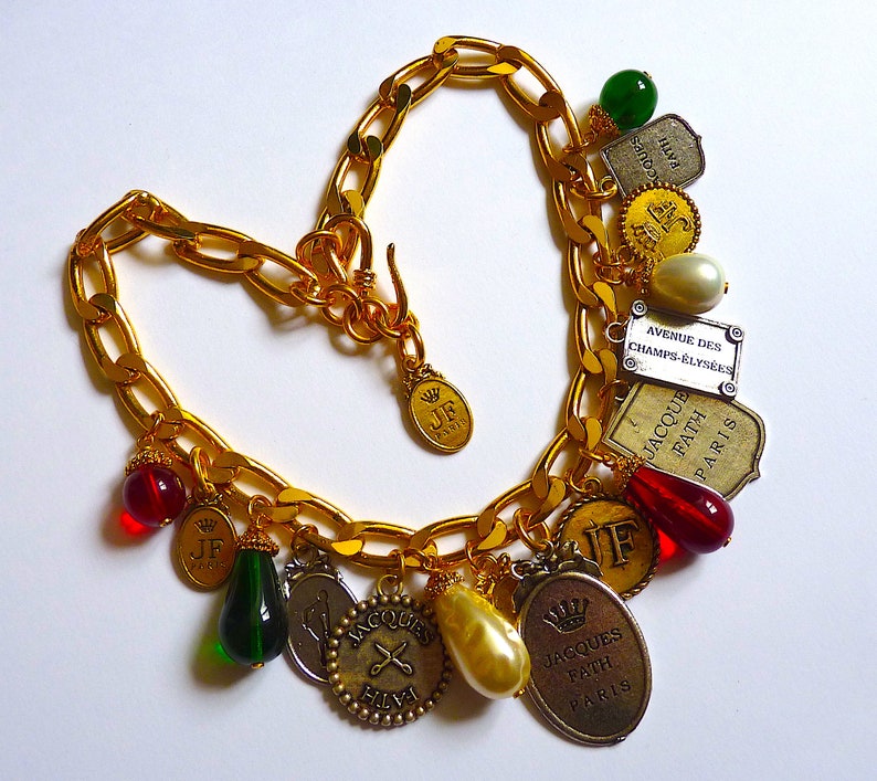 Puede incluir: Un collar de cadena dorada con m&uacute;ltiples dijes, incluyendo cuentas de vidrio rojas y verdes, dijes dorados con el texto "JF Paris" y "Jacques Fath Paris", y un dije plateado con el texto "Avenue des Champs-&Eacute;lys&eacute;es".
