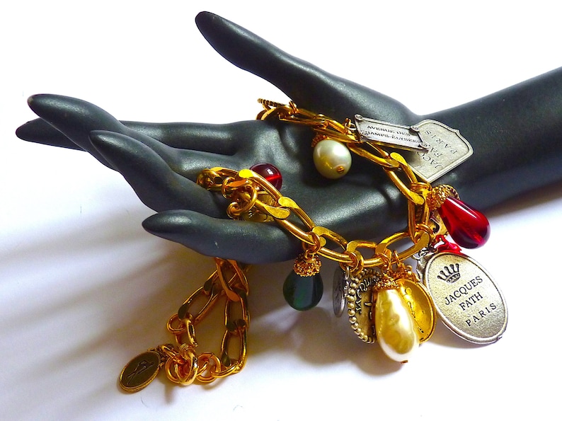 Puede incluir: Un brazalete de cadena dorada con m&uacute;ltiples dijes, incluyendo una cuenta de vidrio roja y verde, una perla y un dije de plata con el texto "JACQUES FATH PARIS".