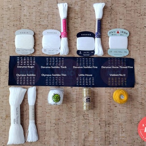 Może przedstawiać: Zestaw białych i ż&oacute;łtych szpulek i kłębk&oacute;w nici do haftu, w tym Daruma Kogin, Daruma Sashiko Thick, Daruma Sashiko Fine, Daruma Home Thread Fine, Olympus Sashiko, Olympus Sashiko Thin, Little House, Valdani No.12 i Sashiko Thread. Szpulki i kłębki są ułożone na powierzchni korkowej z czarną i białą etykietą, na kt&oacute;rej widnieje napis "Daruma Kogin, Daruma Sashiko Thick, Daruma Sashiko Fine, Daruma Home Thread Fine, Olympus Sashiko, Olympus Sashiko Thin, Little House, Valdani No.12".
