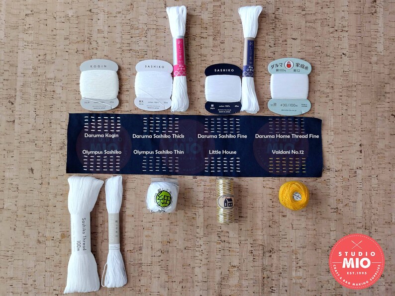 Może przedstawiać: Zestaw białych i ż&oacute;łtych szpulek i kłębk&oacute;w nici do haftu, w tym Daruma Kogin, Daruma Sashiko Thick, Daruma Sashiko Fine, Daruma Home Thread Fine, Olympus Sashiko, Olympus Sashiko Thin, Little House, Valdani No.12 i Sashiko Thread. Szpulki i kłębki są ułożone na powierzchni korkowej z czarną i białą etykietą, na kt&oacute;rej widnieje napis "Daruma Kogin, Daruma Sashiko Thick, Daruma Sashiko Fine, Daruma Home Thread Fine, Olympus Sashiko, Olympus Sashiko Thin, Little House, Valdani No.12".