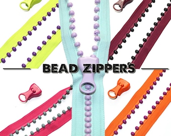 Puede incluir: Surtido de cremalleras con cuentas de colores con cuentas y tiradores de cremallera de diferentes colores. El texto "BEAD ZIPPERS" se muestra en el centro de la imagen.