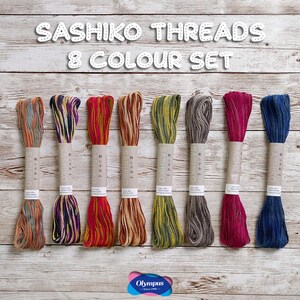 May include: A set of eight different coloured Sashiko threads, including orange, purple, red, brown, green, grey, pink, and blue. The threads are packaged with paper labels and are displayed on a light wood-grain surface. The text "Sashiko Threads 8 Colour Set" is at the top.