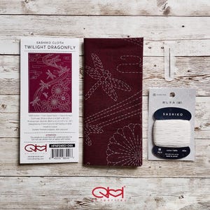 May include: A Sashiko cloth kit with a burgundy dragonfly design, a needle, and white thread. The kit includes a printed fabric piece, a spool of white thread, and a needle. The fabric measures approximately 12 x 16 inches.